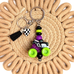 May include: A novelty keychain featuring a miniature monster truck design with green wheels, purple body, and black accents. The keychain includes a black tassel, a checkered heart charm, and a silver keyring. The background is a beige woven coaster.