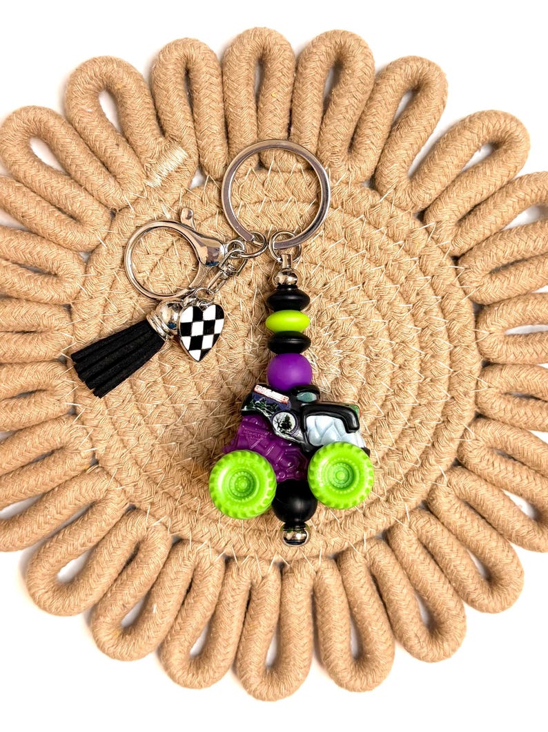 May include: A novelty keychain featuring a miniature monster truck design with green wheels, purple body, and black accents. The keychain includes a black tassel, a checkered heart charm, and a silver keyring. The background is a beige woven coaster.