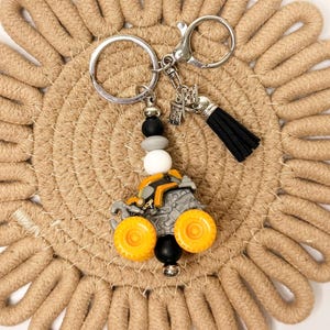 May include: A keychain featuring a miniature orange and gray construction vehicle toy, accented with round black, gray, and white silicone beads, a silver-toned metal lobster clasp, a small silver charm, and a black faux leather tassel.