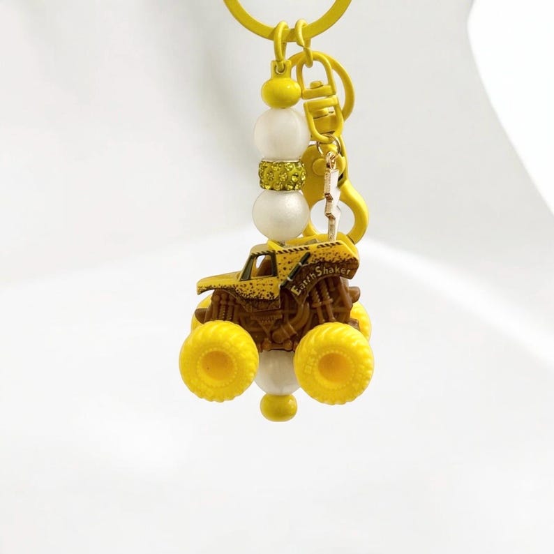 May include: A yellow and brown monster truck keyring with the text "Earth Shaker". The keyring includes white and gold beads and a yellow clasp. The truck has large yellow wheels and a detailed design.