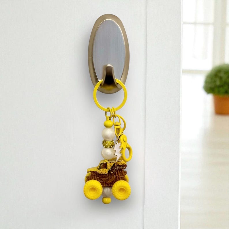 May include: A novelty monster truck keyring in yellow and brown, with a beaded section and a lightning bolt charm. The keyring is suspended from a silver hook on a white door. The truck has yellow wheels and the text "Earth Shaker".