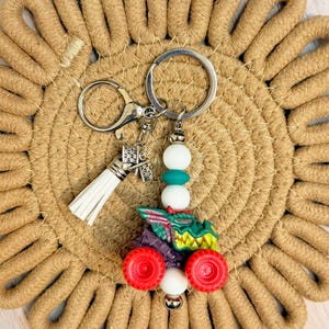 May include: A silver keychain with a white tassel and a charm shaped like a building. The keychain features a tractor charm with red wheels, green and purple details, and white and teal beads.