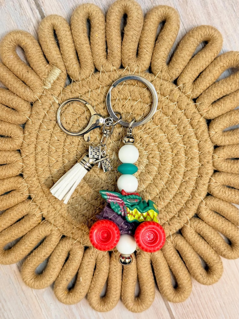 May include: A silver keychain with a white tassel and a charm shaped like a building. The keychain features a tractor charm with red wheels, green and purple details, and white and teal beads.