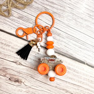 May include: An orange keychain featuring a monster truck charm with the word "ZOMBIE". The keychain includes a black tassel, a lightning bolt charm, and orange and white beads. It is attached to a clasp and a key ring.