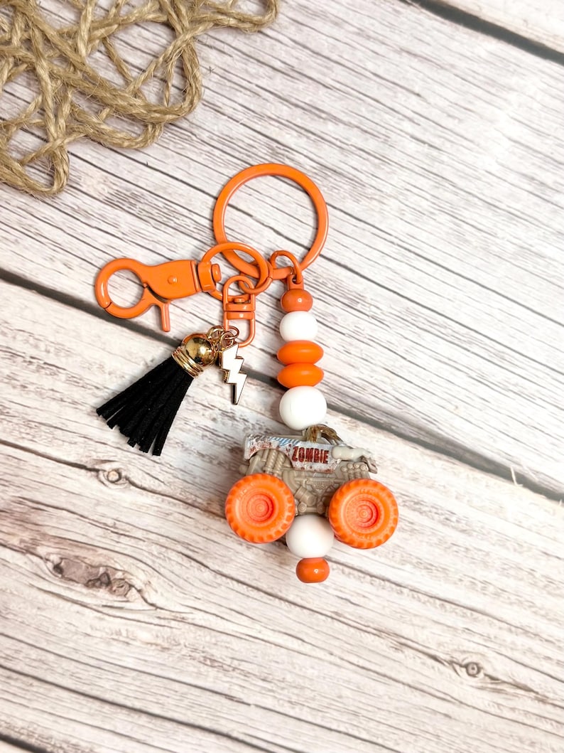 May include: An orange keychain featuring a monster truck charm with the word "ZOMBIE". The keychain includes a black tassel, a lightning bolt charm, and orange and white beads. It is attached to a clasp and a key ring.