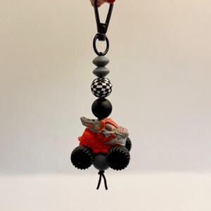 May include: A handmade keychain featuring a small, orange and gray monster truck toy with black wheels. The keychain has a black clasp and is strung with gray, black, and checkered beads. The toy truck is the focal point of the accessory.