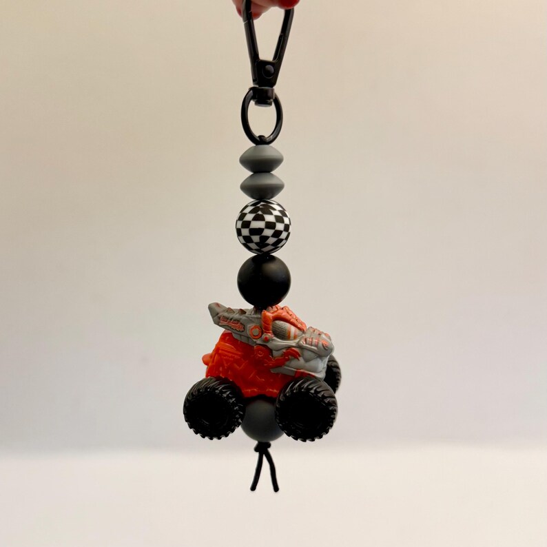 May include: A handmade keychain featuring a small, orange and gray monster truck toy with black wheels. The keychain has a black clasp and is strung with gray, black, and checkered beads. The toy truck is the focal point of the accessory.