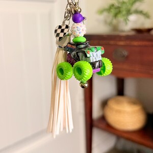 May include: A keychain featuring a miniature monster truck with neon green wheels and a black and green body. The keychain includes a tassel, a heart-shaped charm with a checkered pattern, and decorative beads. The truck is approximately 2 inches long.
