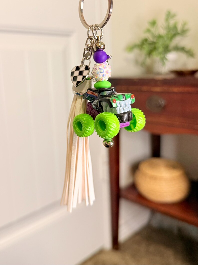 May include: A keychain featuring a miniature monster truck with neon green wheels and a black and green body. The keychain includes a tassel, a heart-shaped charm with a checkered pattern, and decorative beads. The truck is approximately 2 inches long.