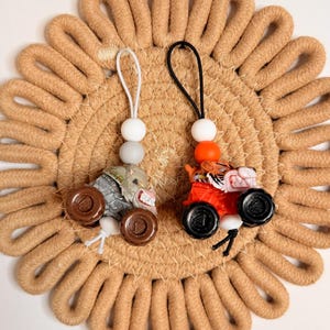 May include: Two miniature toy monster trucks, one gray and one red, each with a beaded charm. The gray truck has brown wheels and a white charm. The red truck has black wheels and an orange charm. They rest on a tan, circular woven mat.