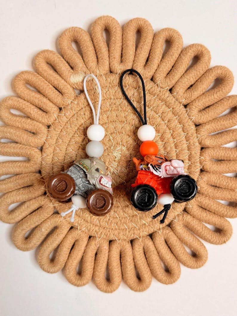 May include: Two miniature toy monster trucks, one gray and one red, each with a beaded charm. The gray truck has brown wheels and a white charm. The red truck has black wheels and an orange charm. They rest on a tan, circular woven mat.