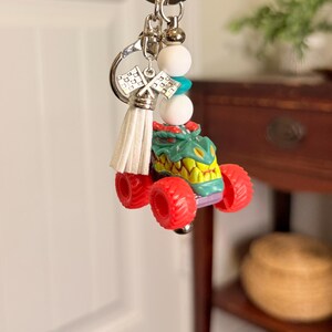 May include: A monster truck keychain with a green and yellow body, red wheels, and a silver key ring. The keychain includes white and turquoise beads, a white tassel, and silver racing flags.