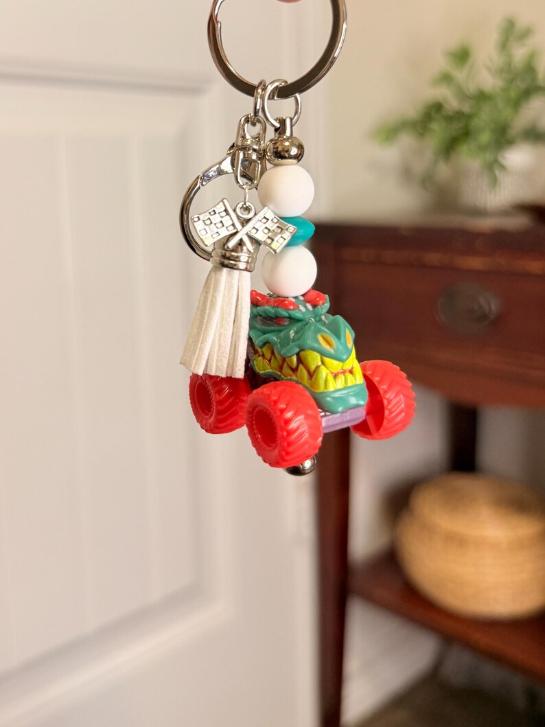 May include: A monster truck keychain with a green and yellow body, red wheels, and a silver key ring. The keychain includes white and turquoise beads, a white tassel, and silver racing flags.