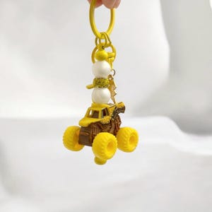 May include: Yellow monster truck keyring with a snowman charm. The truck has large yellow wheels and a brown chassis. The snowman has white spheres for the body and a yellow scarf. A gold lightning bolt charm hangs from the keyring.