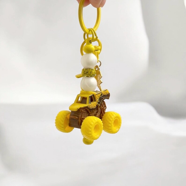 May include: Yellow monster truck keyring with a snowman charm. The truck has large yellow wheels and a brown chassis. The snowman has white spheres for the body and a yellow scarf. A gold lightning bolt charm hangs from the keyring.