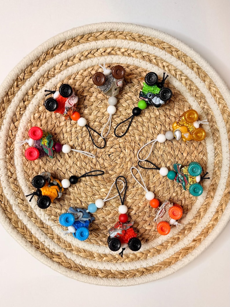 May include: A collection of colorful toy monster truck pacifier clips. Each clip features a miniature truck with vibrant wheels and a beaded chain. The trucks are in various colors, including red, blue, orange, and green.