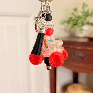 May include: A novelty keychain featuring a miniature clear race car with red wheels. The keychain includes a black tassel, silver-tone metal accents, and colorful beads. The car has a blue seat and is attached to a silver key ring.
