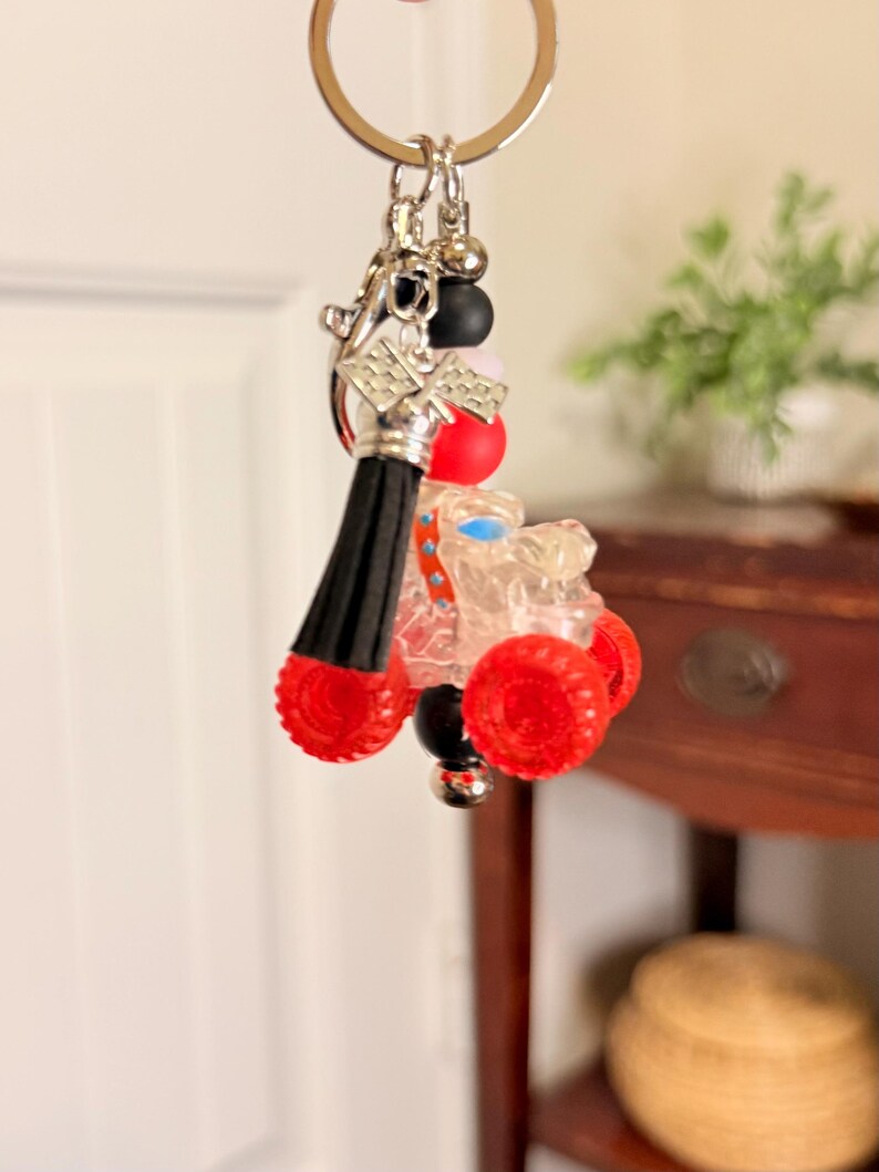 May include: A novelty keychain featuring a miniature clear race car with red wheels. The keychain includes a black tassel, silver-tone metal accents, and colorful beads. The car has a blue seat and is attached to a silver key ring.