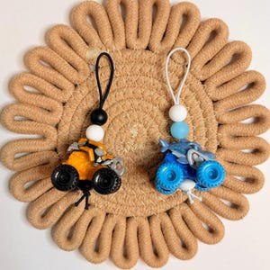 May include: Two toy monster trucks, one orange and one blue, with black wheels and beaded lanyards. They rest on a round, tan woven coaster. The orange truck has a black and white bead, and the blue truck has a white and blue bead.