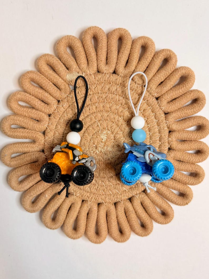 May include: Two toy monster trucks, one orange and one blue, with black wheels and beaded lanyards. They rest on a round, tan woven coaster. The orange truck has a black and white bead, and the blue truck has a white and blue bead.