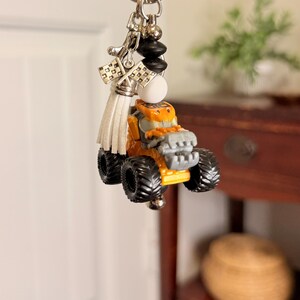 K&ouml;nnte beinhalten: Ein Schl&uuml;sselanh&auml;nger mit einem kleinen orangefarbenen und grauen Monstertruck-Anh&auml;nger. Der Schl&uuml;sselanh&auml;nger hat einen silbernen Ring, schwarze und wei&szlig;e Perlen, eine wei&szlig;e Quaste und karierte Flaggenanh&auml;nger. Der Truck hat gro&szlig;e schwarze Reifen.