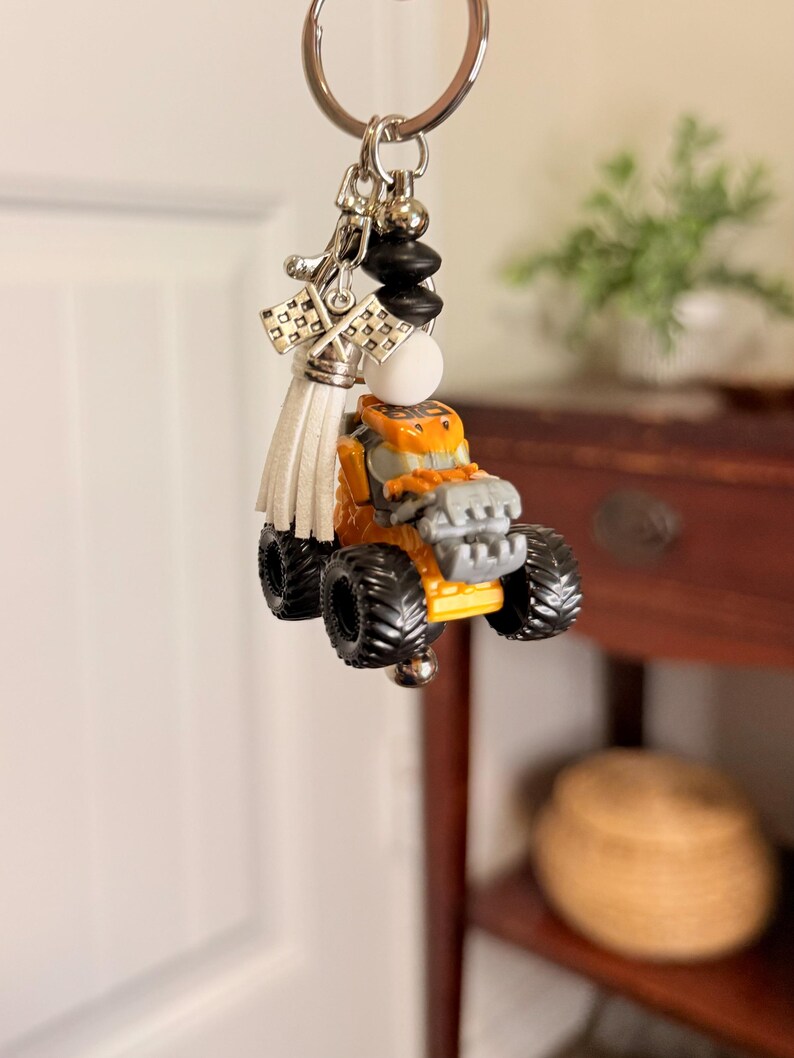 K&ouml;nnte beinhalten: Ein Schl&uuml;sselanh&auml;nger mit einem kleinen orangefarbenen und grauen Monstertruck-Anh&auml;nger. Der Schl&uuml;sselanh&auml;nger hat einen silbernen Ring, schwarze und wei&szlig;e Perlen, eine wei&szlig;e Quaste und karierte Flaggenanh&auml;nger. Der Truck hat gro&szlig;e schwarze Reifen.
