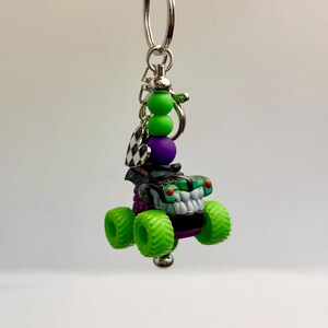 May include: A monster truck keyring with a silver ring. The truck is black and green with a white grill and green tyres. The keyring also features green and purple beads and a checkered flag charm.