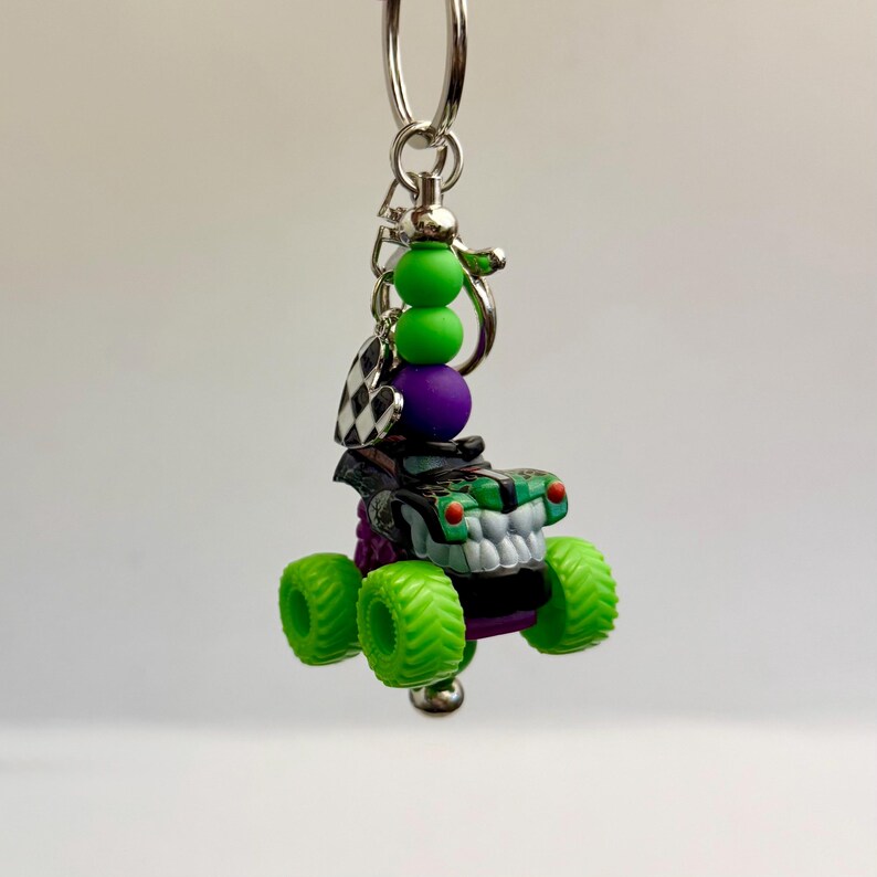 May include: A monster truck keyring with a silver ring. The truck is black and green with a white grill and green tyres. The keyring also features green and purple beads and a checkered flag charm.