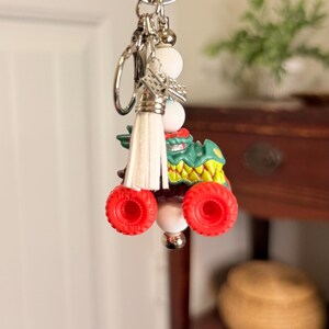 May include: A novelty keychain featuring a green monster truck with red wheels and a white tassel. The keychain includes white beads and a silver keyring. The monster truck has yellow and green accents.