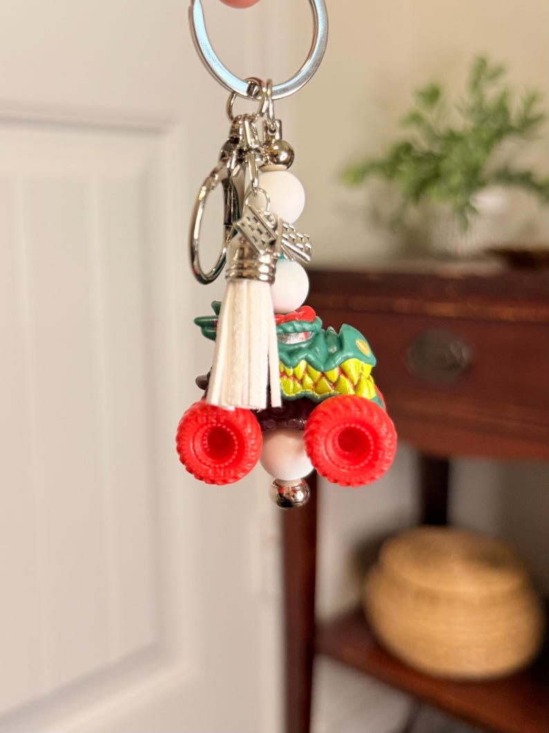 May include: A novelty keychain featuring a green monster truck with red wheels and a white tassel. The keychain includes white beads and a silver keyring. The monster truck has yellow and green accents.