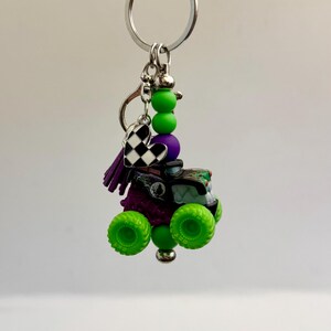 May include: A monster truck keyring with green wheels, purple detailing, and a black and white chequered heart charm. The keyring also features green and purple beads, and a silver-tone ring.