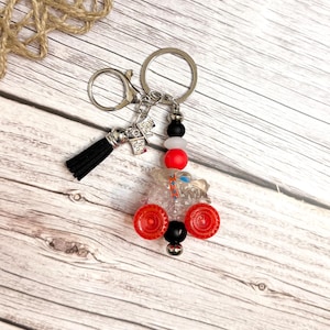 May include: A handmade keychain featuring a clear, miniature tractor with red wheels and black and white beads. The keychain includes a black tassel and a silver clasp and ring. The background is a light wood surface.