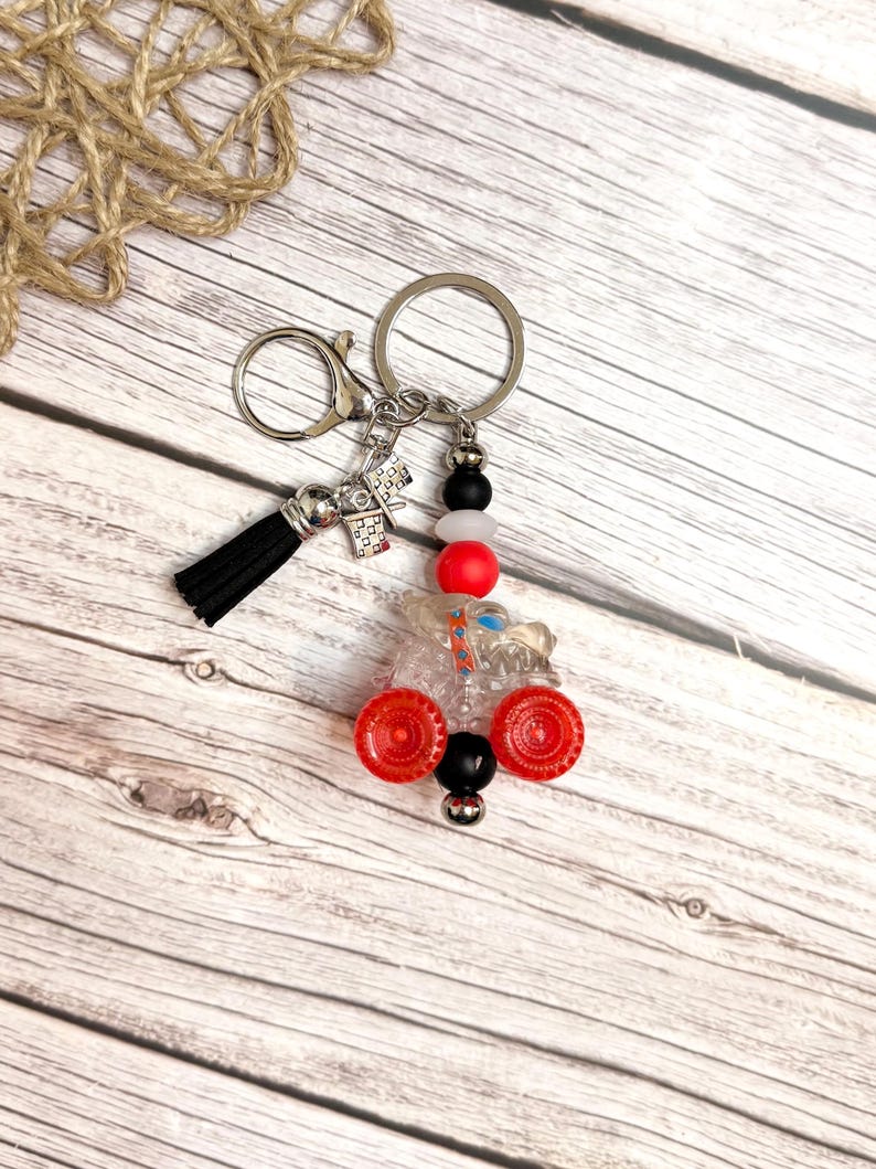 May include: A handmade keychain featuring a clear, miniature tractor with red wheels and black and white beads. The keychain includes a black tassel and a silver clasp and ring. The background is a light wood surface.