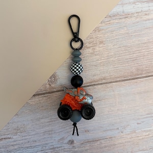 May include: A monster truck keychain with a black clasp, gray and black beads, and an orange and gray truck. The truck has large black tires and a detailed design. The keychain is attached to a black cord.