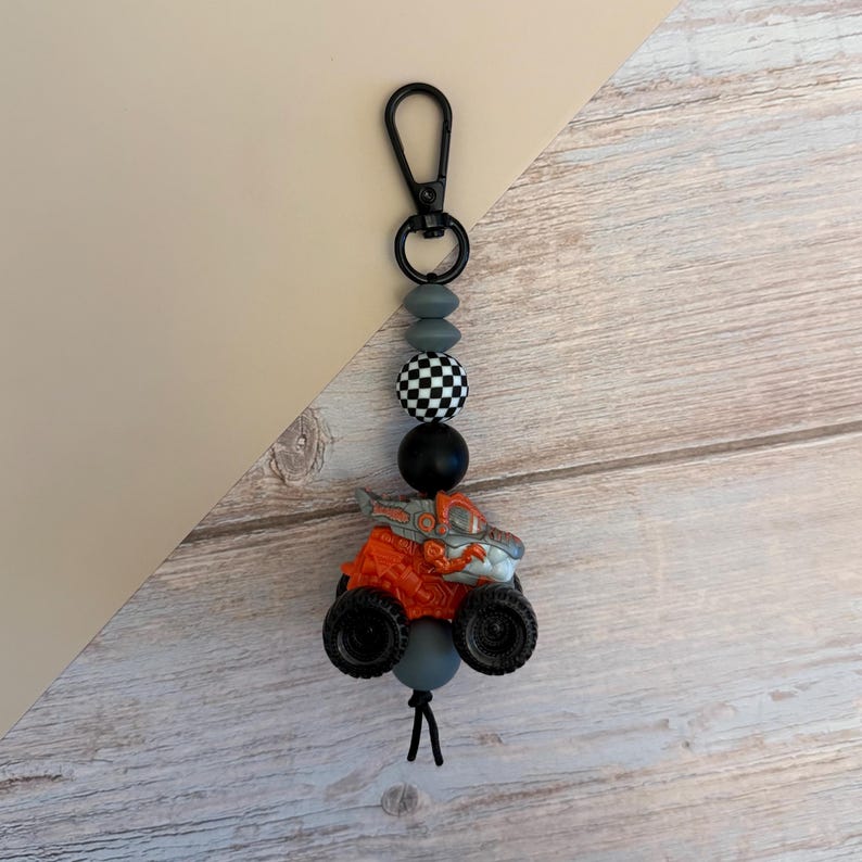 May include: A monster truck keychain with a black clasp, gray and black beads, and an orange and gray truck. The truck has large black tires and a detailed design. The keychain is attached to a black cord.