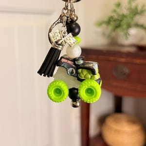 May include: A keychain featuring a miniature monster truck with neon green wheels and accents. The truck has a black and silver body with the words "Grave Digger" visible. The keychain also includes a black tassel, silver charms, and various beads.