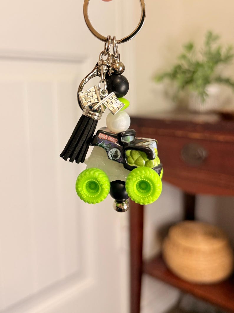 May include: A keychain featuring a miniature monster truck with neon green wheels and accents. The truck has a black and silver body with the words "Grave Digger" visible. The keychain also includes a black tassel, silver charms, and various beads.