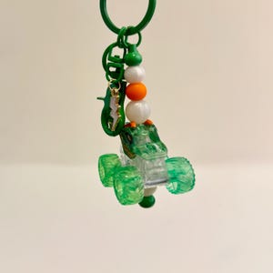 May include: A green and clear monster truck keychain with a green key ring. The keychain features a green monster truck with clear windows and green wheels. The keychain also has a charm of a green alligator and a gold lightning bolt.