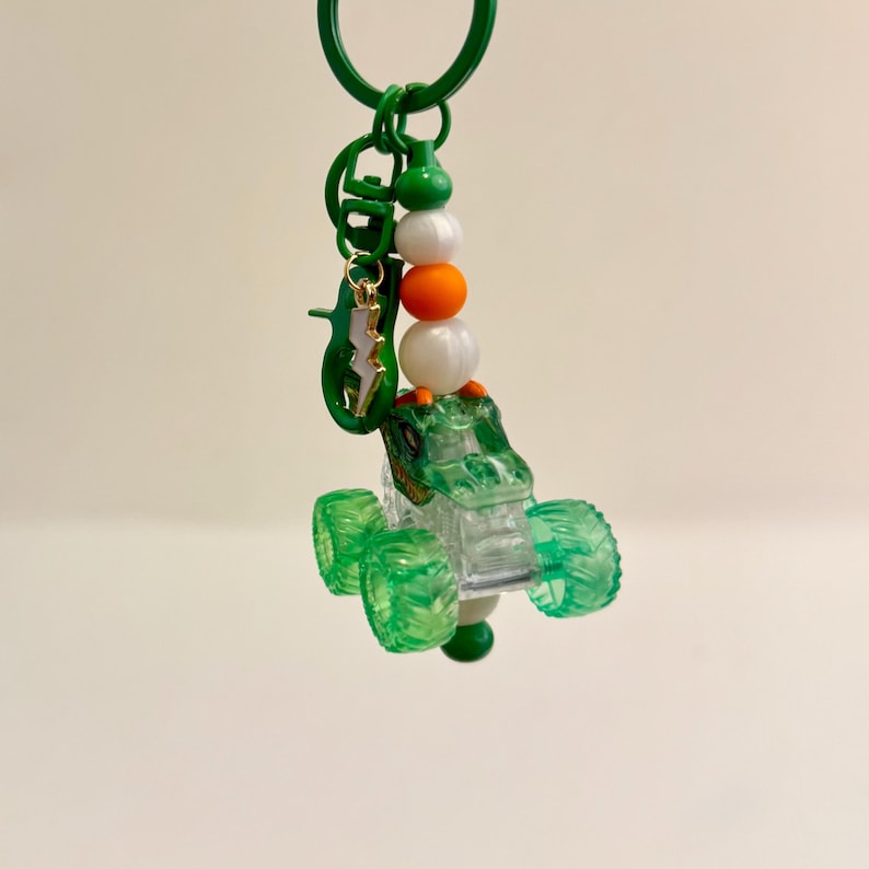 May include: A green and clear monster truck keychain with a green key ring. The keychain features a green monster truck with clear windows and green wheels. The keychain also has a charm of a green alligator and a gold lightning bolt.