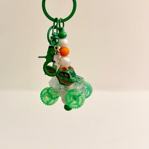 May include: A green monster truck keychain with a green alligator head and clear wheels. The keychain has a green ring, a green clasp, and a charm of a white and gold lightning bolt. White, orange, and green beads are also attached.