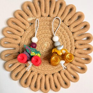 May include: Two miniature monster truck ornaments. One is green, purple, and red, and the other is yellow and silver. Each has a white string with a bead and a loop for hanging. They rest on a tan, circular woven coaster.