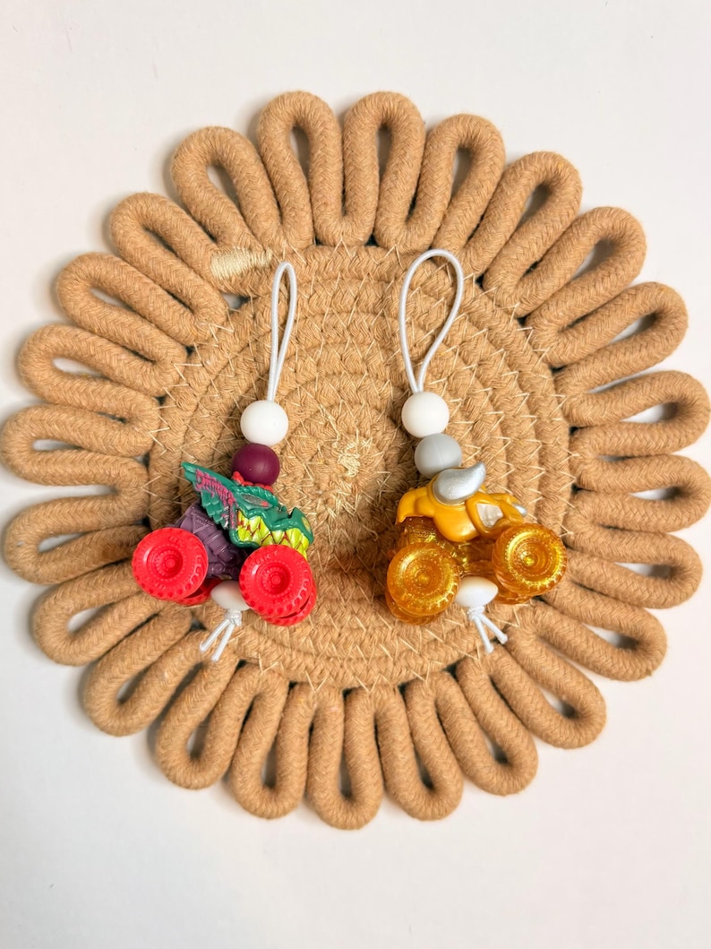 May include: Two miniature monster truck ornaments. One is green, purple, and red, and the other is yellow and silver. Each has a white string with a bead and a loop for hanging. They rest on a tan, circular woven coaster.