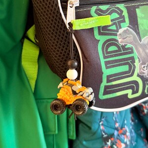 May include: An orange and gray toy ATV with black wheels hangs from a black string with a black and white bead. The ATV is attached to a black backpack with green accents and the word "JURASSIC" in green.