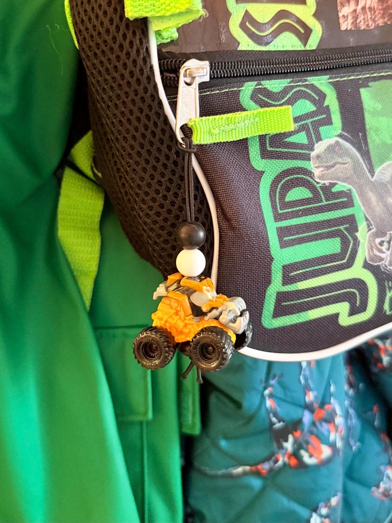 May include: An orange and gray toy ATV with black wheels hangs from a black string with a black and white bead. The ATV is attached to a black backpack with green accents and the word "JURASSIC" in green.