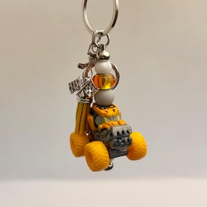 May include: A yellow and grey monster truck keyring with a silver ring. The keyring includes a small tassel, beads, and a chequered flag charm. The truck has large yellow tyres and a detailed design.