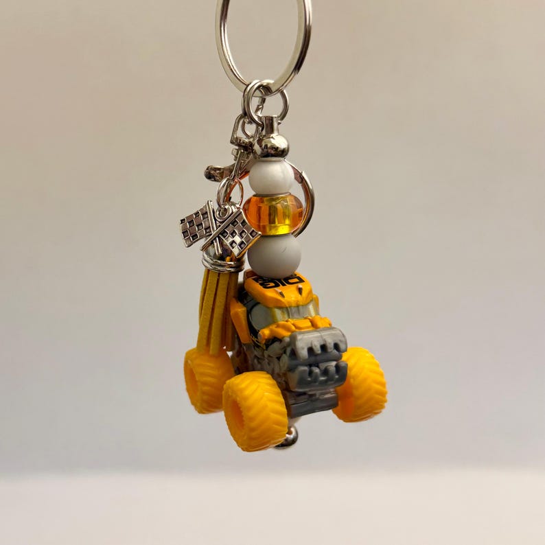 May include: A yellow and grey monster truck keyring with a silver ring. The keyring includes a small tassel, beads, and a chequered flag charm. The truck has large yellow tyres and a detailed design.