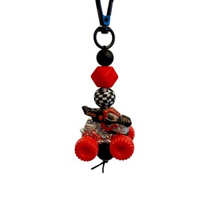 May include: A red and black monster truck charm with a checkered bead, red cube bead, and black beads. The charm is attached to a black clip. The monster truck has red wheels and a silver engine.