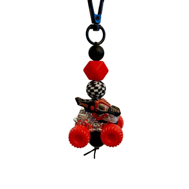 May include: A red and black monster truck charm with a checkered bead, red cube bead, and black beads. The charm is attached to a black clip. The monster truck has red wheels and a silver engine.