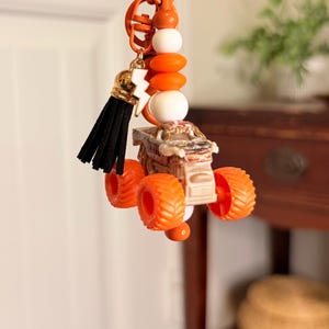 May include: Orange keyring featuring a miniature monster truck with orange wheels. The keyring includes orange, white, and gold beads, a black tassel, and a gold lightning bolt charm. The keyring is attached to an orange metal ring.
