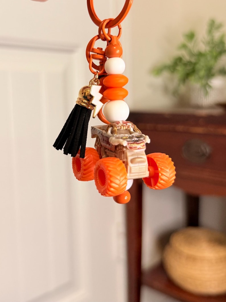 May include: Orange keyring featuring a miniature monster truck with orange wheels. The keyring includes orange, white, and gold beads, a black tassel, and a gold lightning bolt charm. The keyring is attached to an orange metal ring.